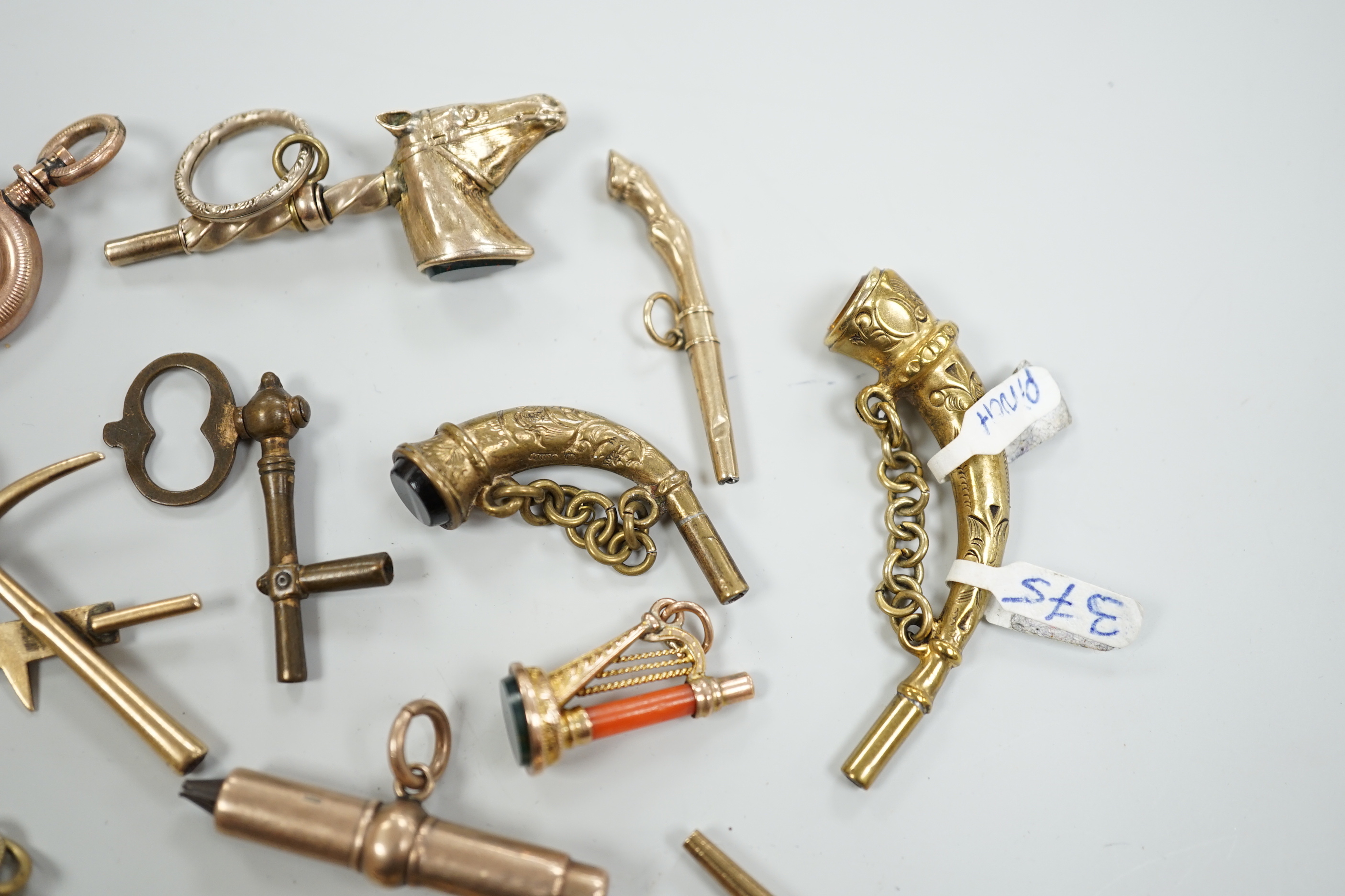 Eighteen assorted mainly 19th century base or yellow metal overlaid novelty watch keys, including coral and bloodstone set harp, pistol, rifle, cornucopia, bellows, horses hoof and trade insignia, largest 55mm.